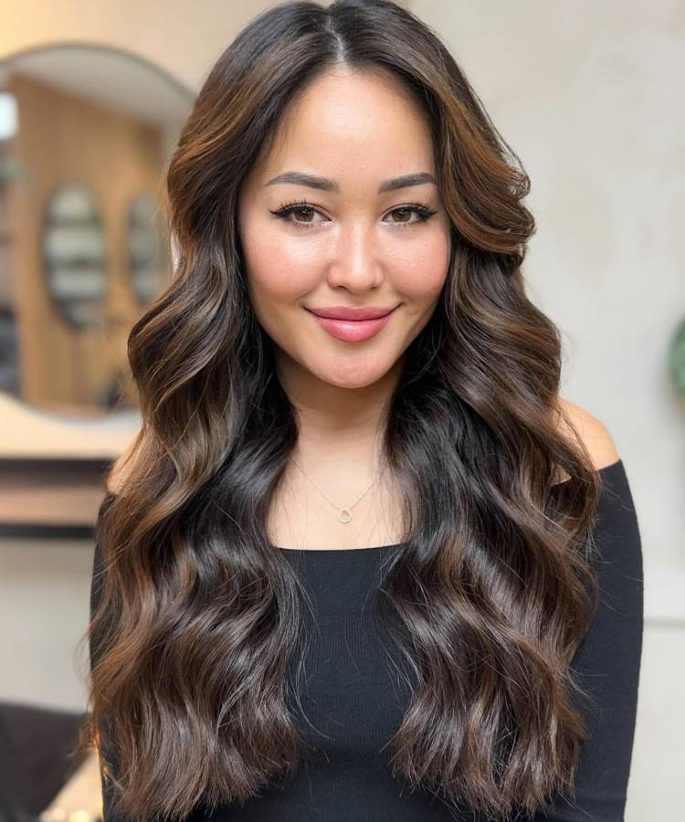 A imagem mostra uma mulher sorrindo levemente, com longos cabelos castanhos iluminados e ondulados. Ela usa uma blusa preta de ombros caídos e um colar delicado. A iluminação destaca seu rosto e o volume do cabelo, com um fundo desfocado de ambiente interno.







