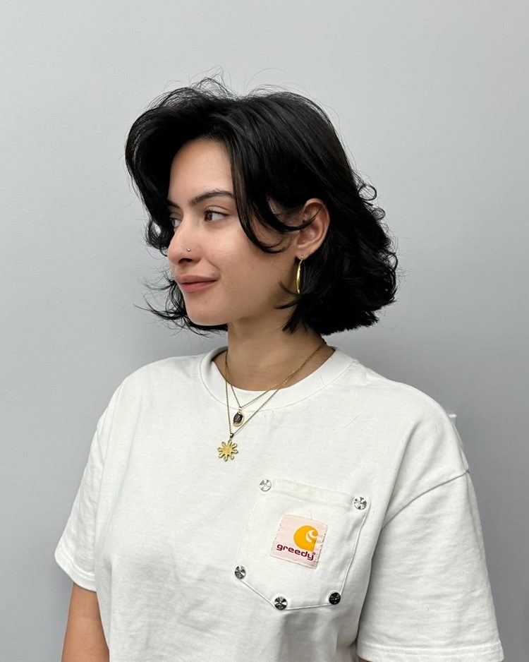 Mulher de perfil com cabelo curto e ondulado, usando camiseta branca com bolso decorado. Ela tem brincos de argola dourados e colares delicados. A expressão é tranquila, destacando um estilo casual e moderno contra um fundo cinza.


