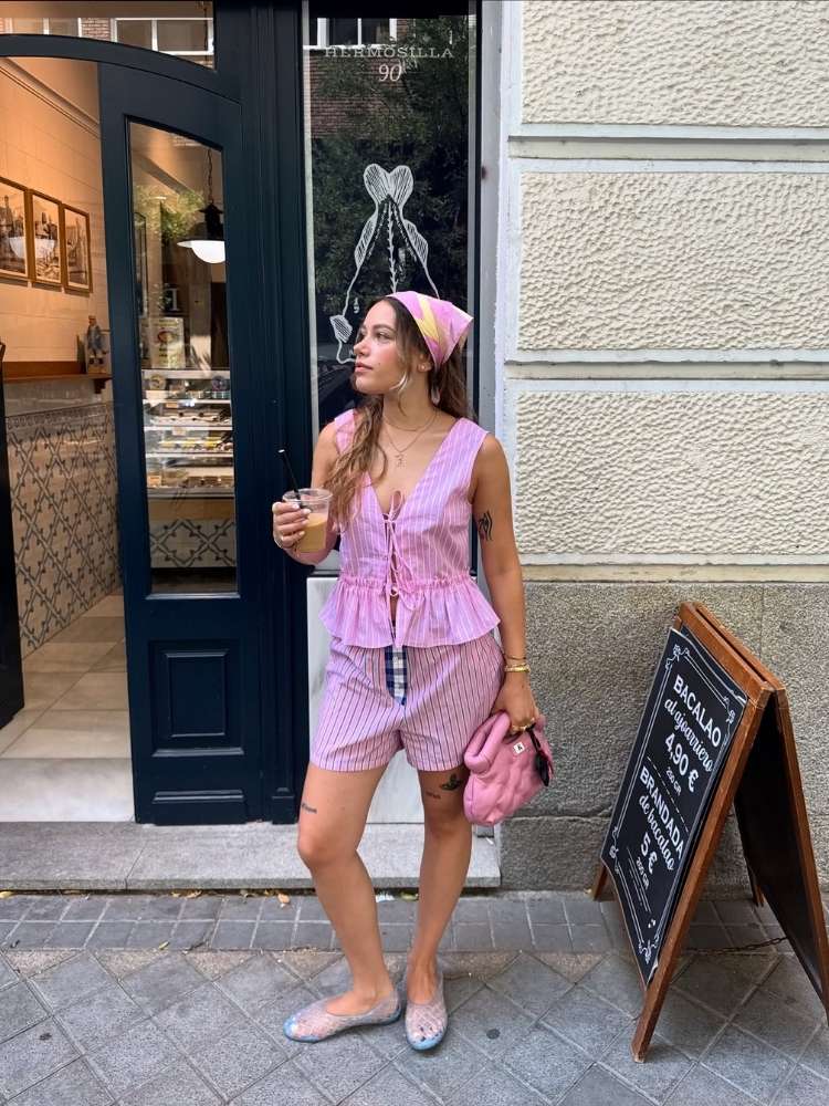 Mulher de pele clara com blusa rosa listrada de amarração, shorts rosa listrado, sapatilha transparente e bandana rosa 
