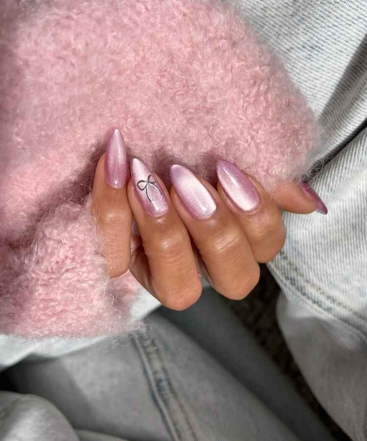 Unhas longas com esmalte rosa perolado e detalhe de laço preto em uma delas. O fundo é tecido suave, também rosa, destacando o brilho sutil das unhas. O visual é delicado, sofisticado e feminino, com foco nas texturas.