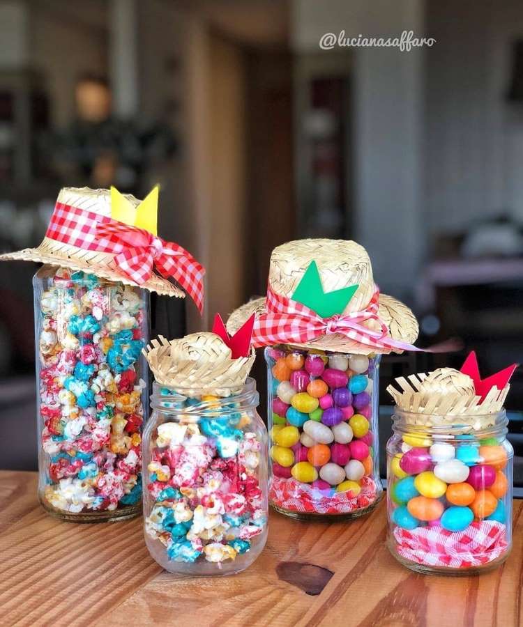 Quatro potes de vidro decorativos, três com pipoca colorida e um com doces variados, todos adornados com chapéus de palha em miniatura e laços xadrez. Perfeitos para lembrancinhas ou centro de mesa juninos.