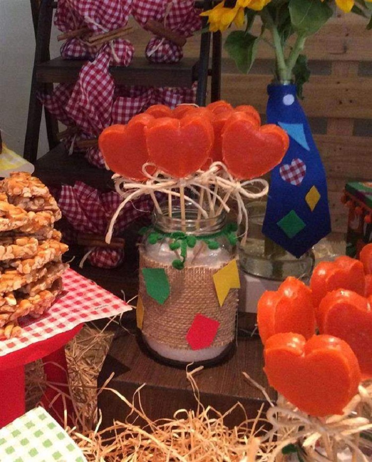 A imagem representa doce de abóbora em formato de coração dentro de pote de vidro decorado com bandeirinhas, sob mesa cheia de mais doces e decorações