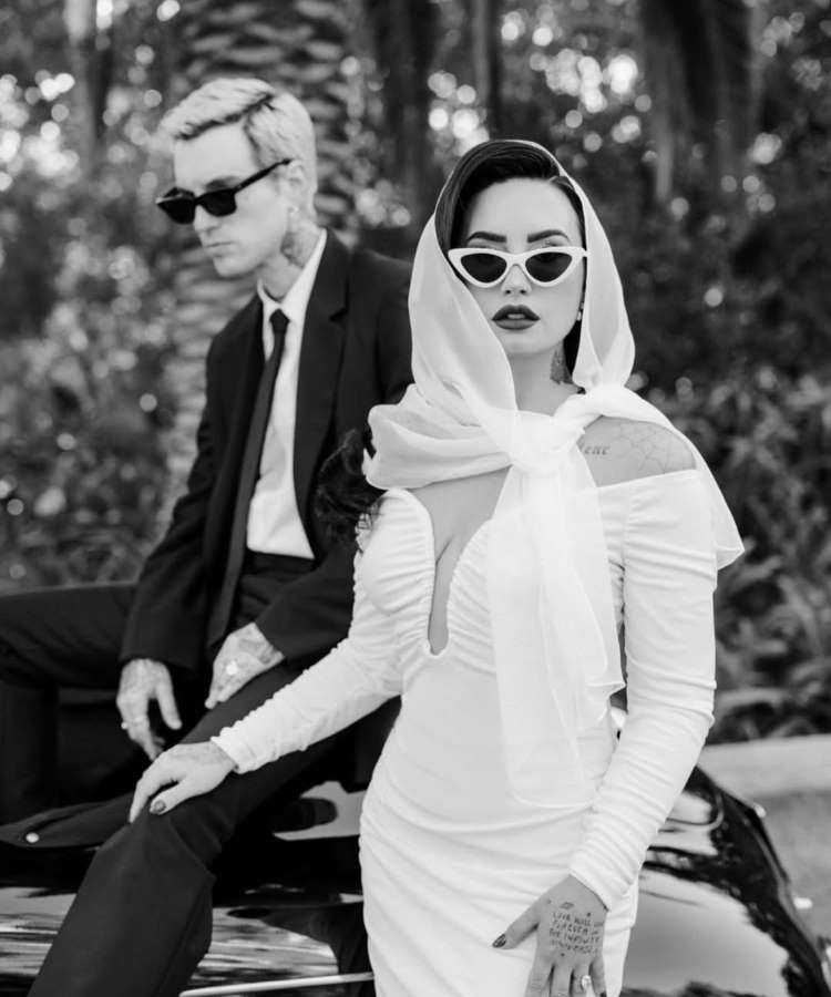 Em preto e branco, Demi Lovato está em primeiro plano, com um lenço na cabeça e óculos de sol brancos, apoiada em um carro. Um homem com cabelo loiro e óculos de sol escuros está sentado na traseira do carro, com palmeiras ao fundo.