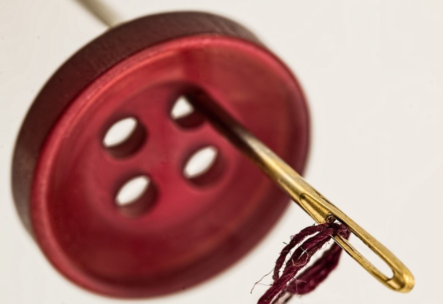 Imagem de um botão vermelho achato de 4 furos atravessado por uma agulha dourada e linha vermelha