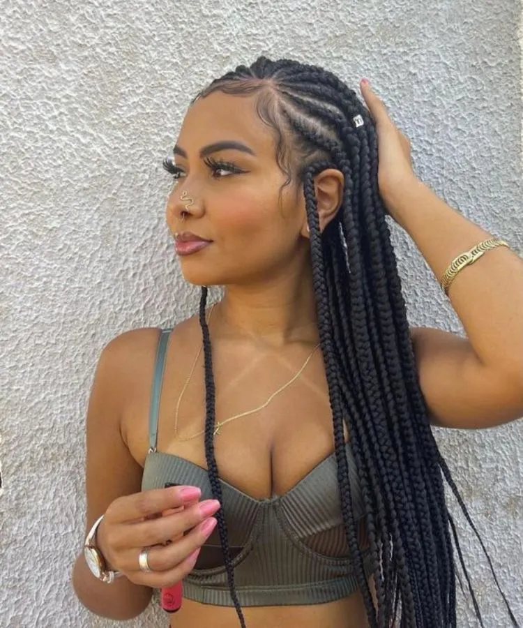 Menina com cabelo trançado, usando top verde-escuro, joias e piercing dourados. Relógio e segurando um produto que parece gloss
