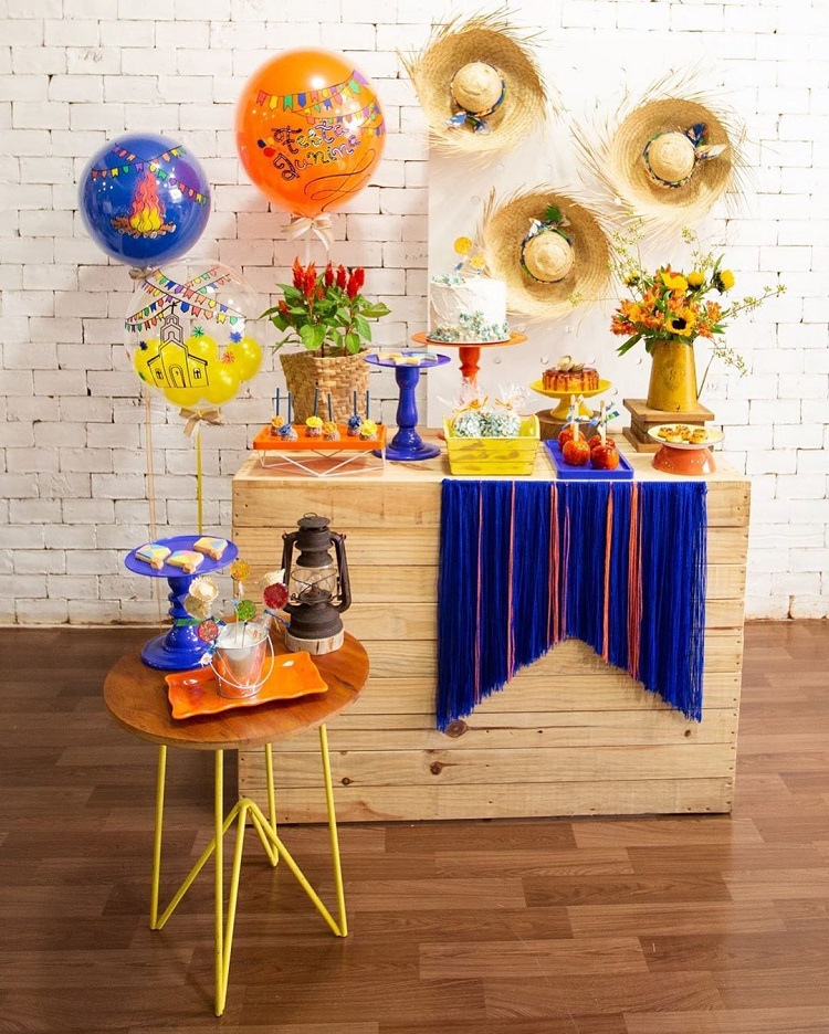 A imagem representa decoração para festa junina com mesas diferentes, em formato de caixote de madeira. Contém doces, balões, chapéus de palha, flores, etc