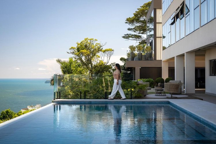 Mulher de pele clara andando ao lado de piscina com borda infinita e mansão atrás