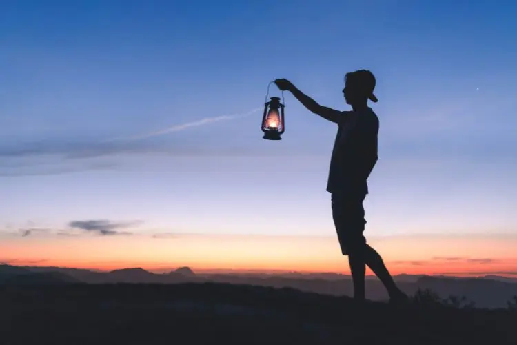 Um jovem segura uma lamparina acesa ao entardecer, com montanhas e o céu colorido de tons alaranjados e azul ao fundo. A silhueta cria uma atmosfera de mistério, reflexão e conexão com a natureza em um momento introspectivo.