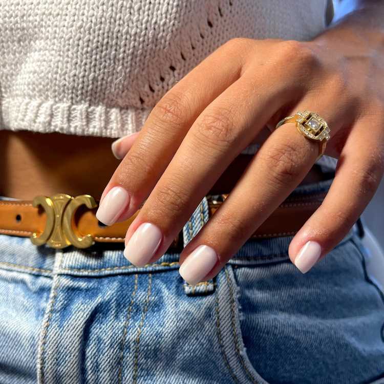 Mão feminina com unhas pintadas em esmalte branco, exibindo anel dourado com pedra brilhante no dedo anelar. Vestindo jeans azul claro com cinto marrom com fivela dourada e top branco de tricô, compondo um visual casual e elegante.