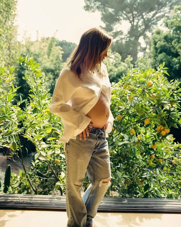 Atris Ashley Tisdale de The Suite Life of Zack & Cody grávida em 2024, usando camisa branca e calça jeans em local com pé de fruta 