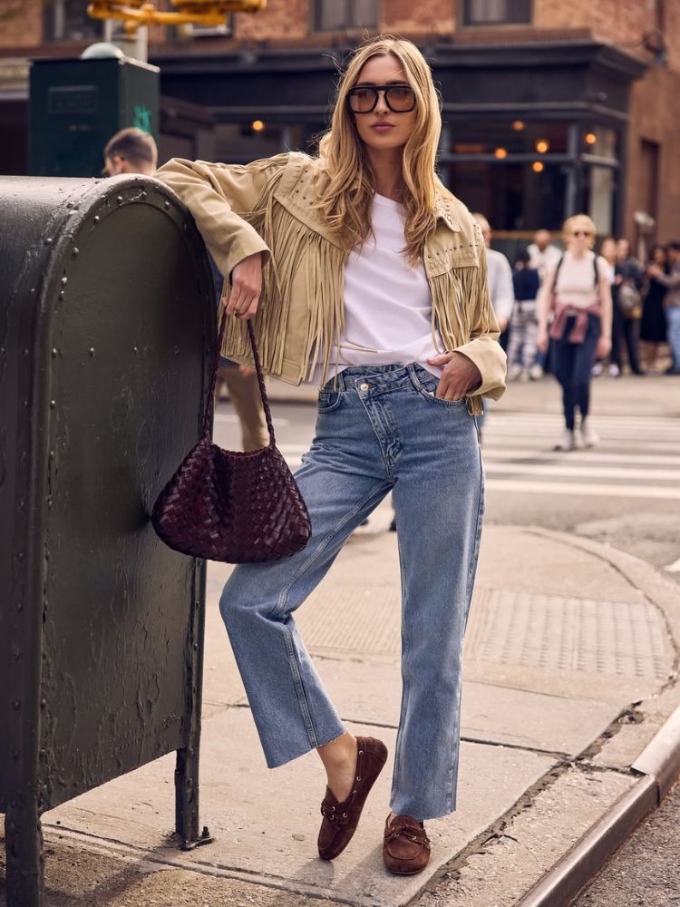 Mulher de pele clara usando camiseta branca, calça jeans, jaqueta creme com franjas e mocassim marrom com rebite 