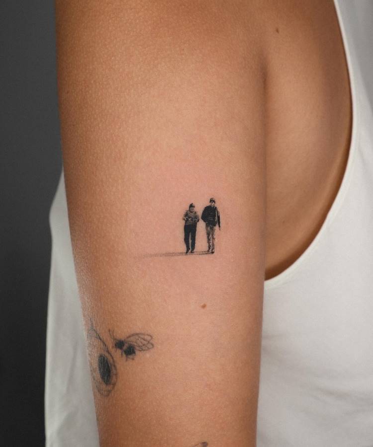 A imagem exibe uma pequena tatuagem minimalista no braço de uma pessoa, retratando duas figuras de corpo inteiro, possivelmente masculinas, caminhando lado a lado. A tatuagem é em preto e cinza, com sombras que dão profundidade. Abaixo, há um esboço de outra tatuagem com abelhas e uma flor.







