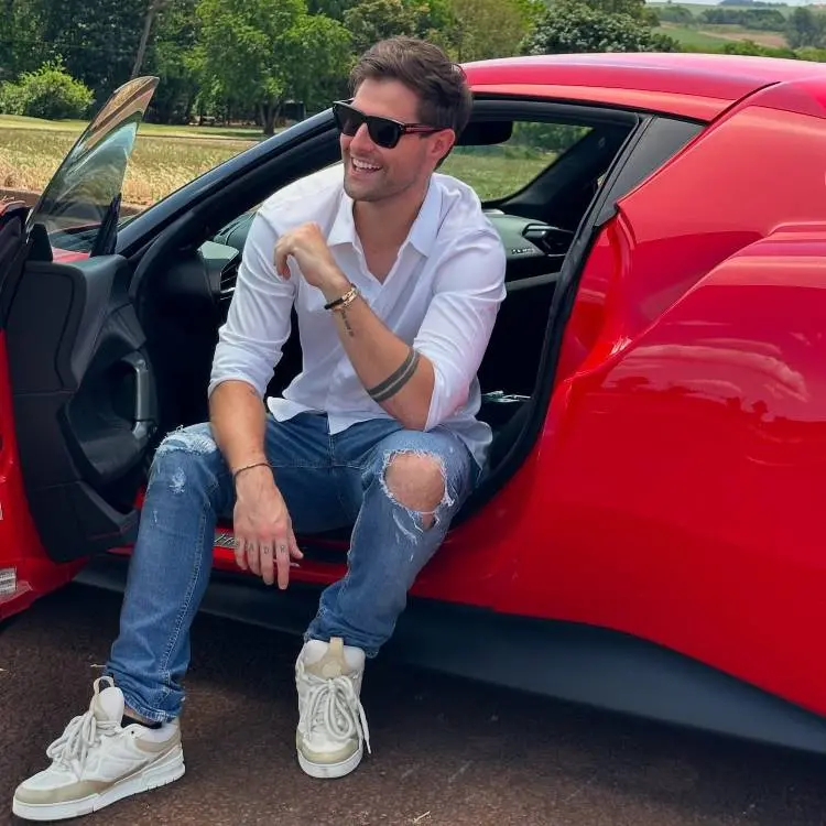 A imagem mostra um homem jovem e sorridente, com óculos escuros e camisa branca, sentado na porta de um carro esportivo vermelho. Ele veste jeans rasgados e tênis brancos, com árvores verdes ao fundo sob um céu claro.







