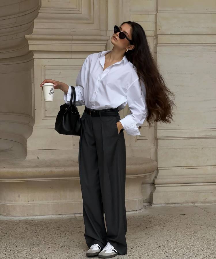 Mulher encostada em parede clássica, veste camisa branca e calça social cinza. Usa óculos escuros retrô, segura um café na mão direita e bolsa preta na esquerda. Cabelos longos soltos, sapatos casuais brancos e cinza completam o look.





