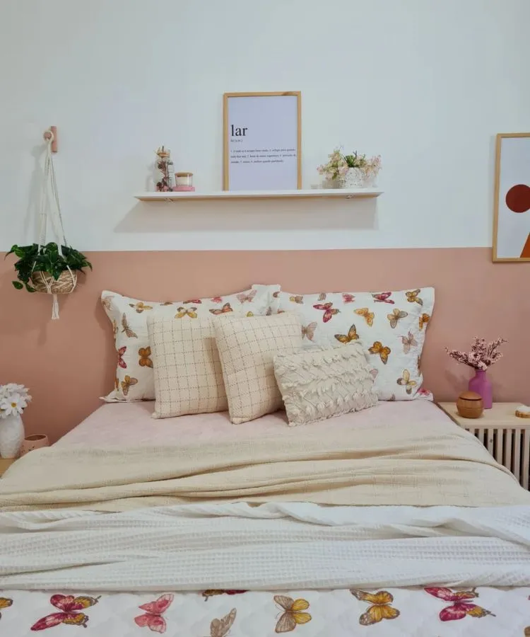 A imagem mostra uma decoração fácil para quarto com uma cama de casal, parede rosa pastel ao fundo, colcha com estampa de borboleta e almofadas variadas. Acima da cabeceira, há uma prateleira branca com vasos de plantas e um quadro com a palavra “lar”. O ambiente é acolhedor e tranquilo, ideal para relaxamento.

