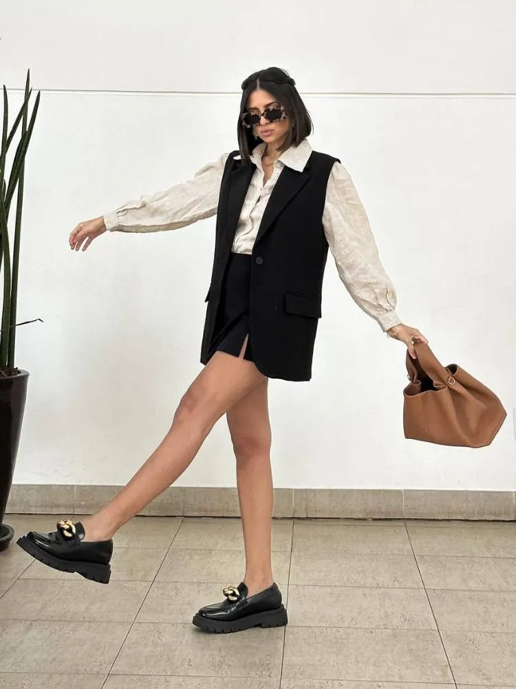 mulher usando camisa de linho, colete e saia pretos e mocassim com salto tratorado