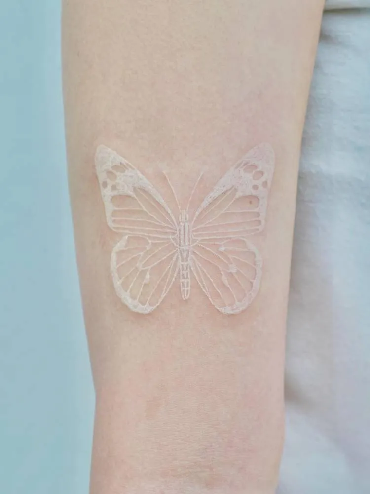 Tatuagem de borboleta com traços finos e um pouco mais grossos na cor branca. Foi feita no braço de uma pessoa de pele clara. 