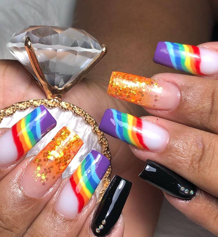 Mão com unhas decoradas coloridas: Polegar: não aparece Indicador: branco com arco-íris Médio: Nude com laranja e glitter Anelar: branco com arco-íris Mínimo Preto