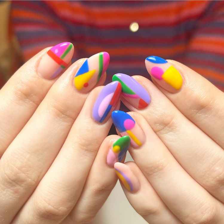 Unhas longas e ovais decoradas com esmalte em tons vibrantes de amarelo, laranja, verde, azul e vermelho, criando um design abstrato e colorido. O fundo escuro contrasta com as cores vivas, destacando o estilo ousado e artístico das unhas.