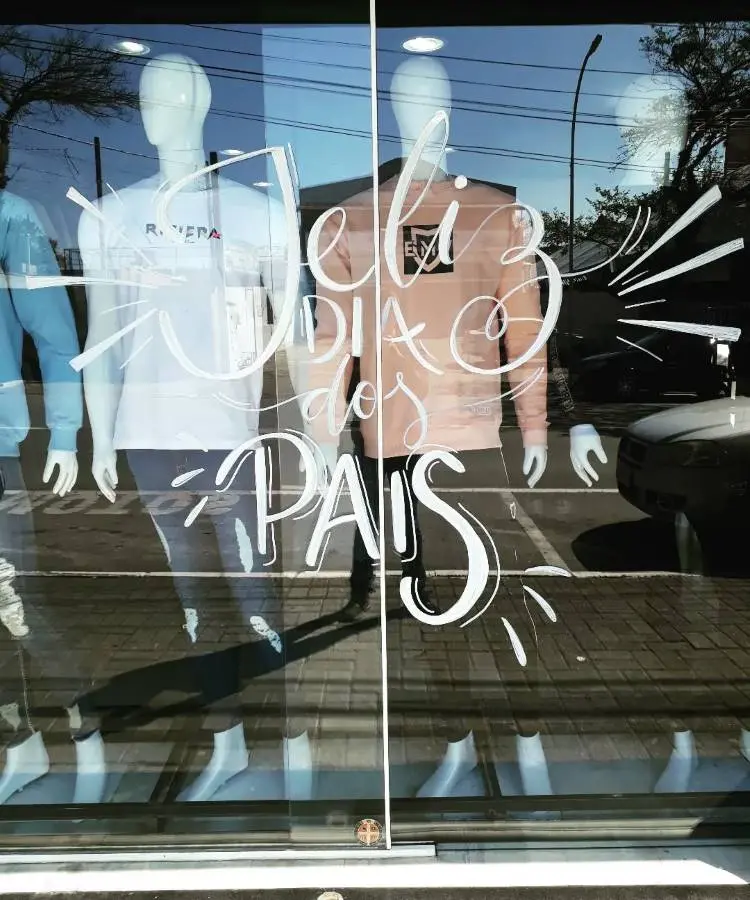 A imagem mostra uma vitrine de loja com dois manequins masculinos exibindo roupas casuais. Na vitrine, está escrito em branco "Feliz Dia dos Pais". Há reflexos do lado de fora, incluindo árvores e carros.