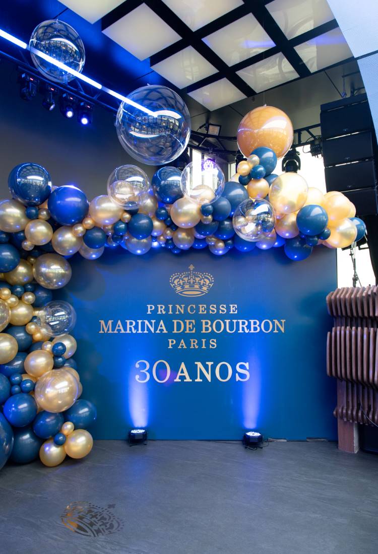 Painel azul com a inscrição "Princesse Marina de Bourbon Paris 30 anos", decorado com balões dourados e azuis. Bolhas transparentes complementam a decoração. Luzes azuis focam o fundo, dando um aspecto festivo e elegante ao cenário comemorativo do evento.