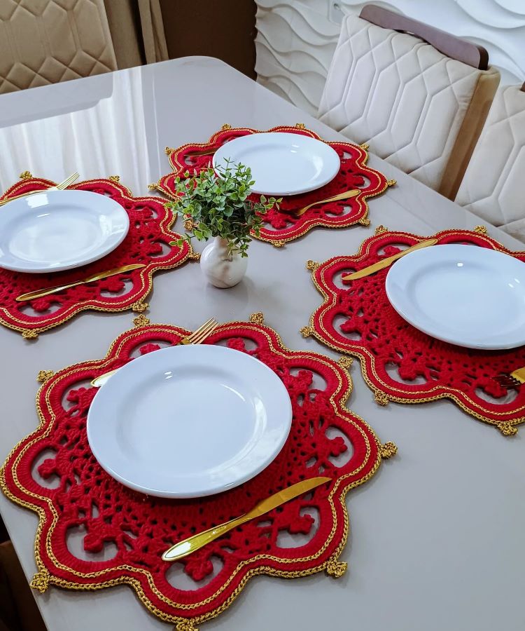 Mesa elegante com sousplats de crochê vermelho e dourado. Louças brancas e talheres dourados. Pequeno vaso com planta verde no centro. Cadeiras acolchoadas ao redor completam a decoração sofisticada.
