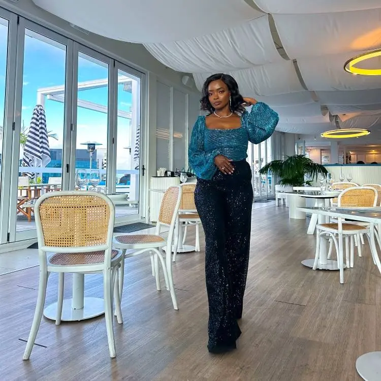 A mulher posa em um restaurante sofisticado, vestindo uma blusa azul brilhante de mangas bufantes e calça preta com glitter. O ambiente iluminado, com teto decorado e cadeiras de madeira, destaca seu visual elegante e contemporâneo.