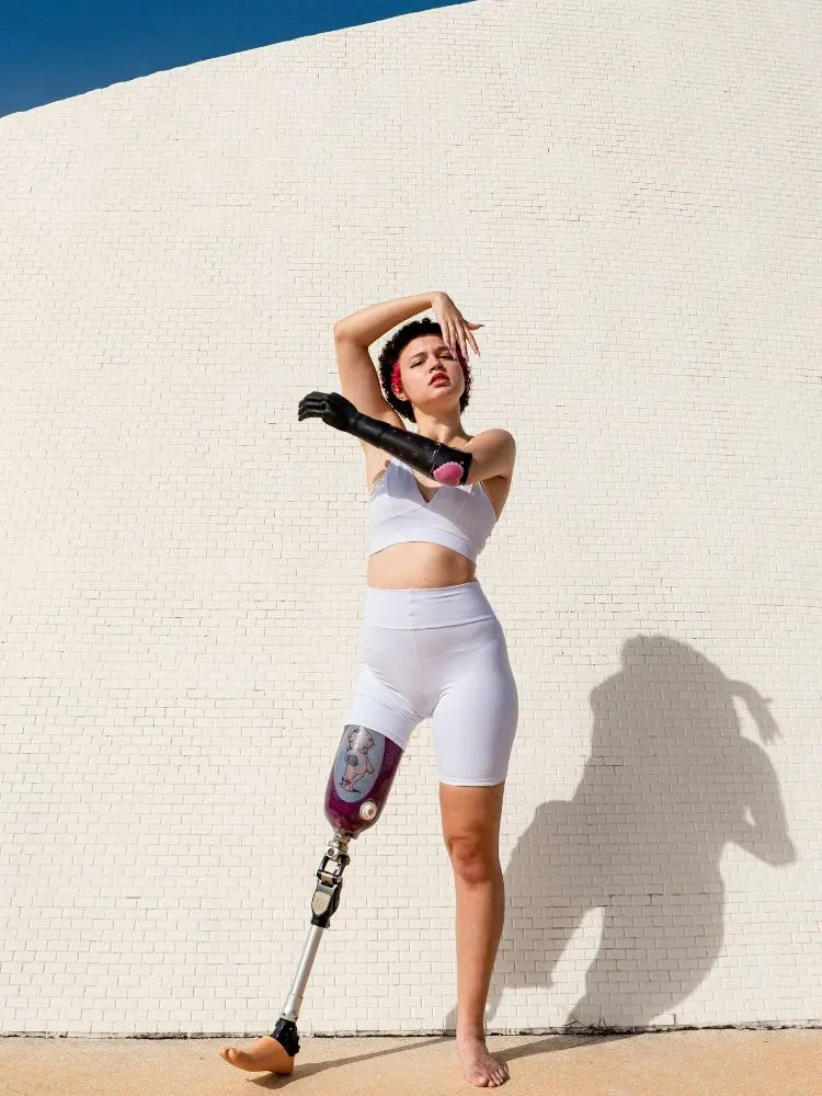 Mulher com deficiência física, braço e perna mecânica, usando conjunto branco de roupa de acadêmia. Ela faz passo de dança em frente à parede clara