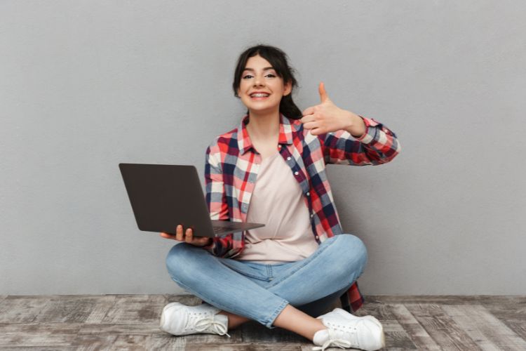 Mulher jovem sorrindo, sentada no chão com as pernas cruzadas, segurando um laptop em uma mão e fazendo sinal de positivo com a outra. Veste camisa xadrez, jeans e tênis, em frente a uma parede cinza.