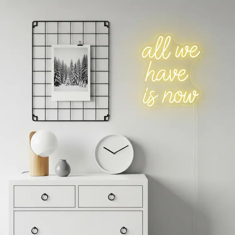 Uma mesa branca com gavetas. Sobre ela, há uma luminária de madeira, um relógio branco, um vaso cinza e, na parede, um neon amarelo diz "all we have is now" ao lado de um quadro de fotos.








