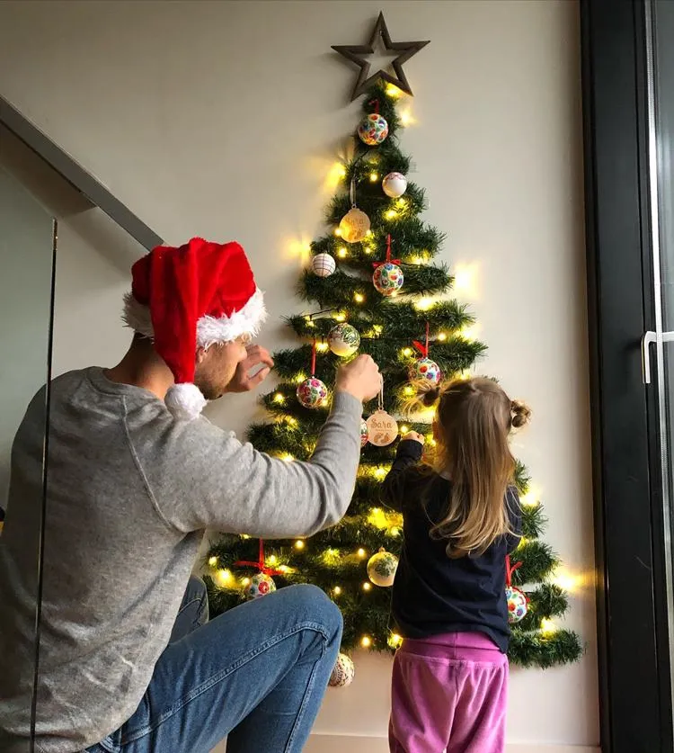 Pai e filha decorando árvore de Natal de parede, feita de festão verde e decorada com bolas natalinas
