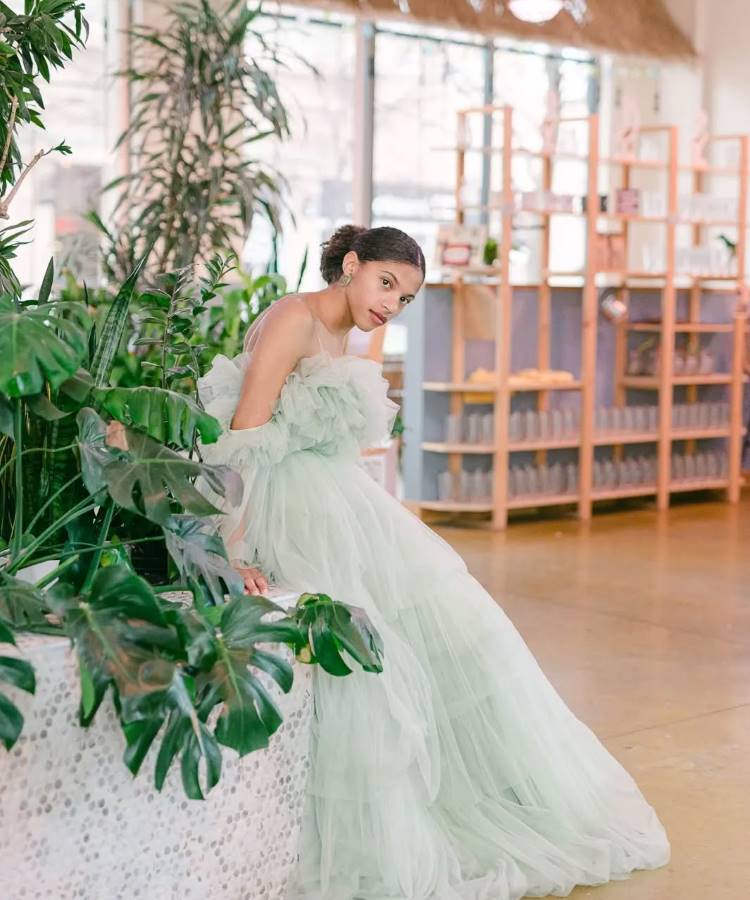Uma noiva de pele morena e penteado preso, vestindo um exuberante vestido de tule verde-água com babados, posa reclinada entre plantas verdes. Ao fundo, prateleiras de madeira com objetos claros e muita luz natural.







