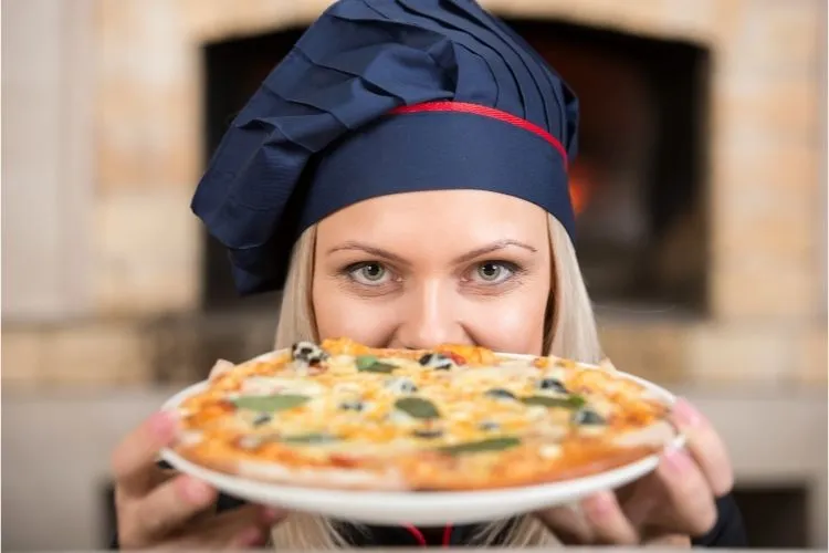 Uma chef loira, vestindo um chapéu de chef azul-marinho com uma fita vermelha, segura uma pizza fresca com azeitonas pretas e manjericão na altura do peito, em frente a um forno a lenha, olhando para a câmera.