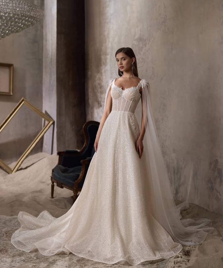 Uma modelo com cabelos escuros, usando um vestido de noiva branco brilhante com espartilho, alças decoradas e uma cauda longa e fluida, posa em um ambiente rústico com uma cadeira escura e molduras douradas.







