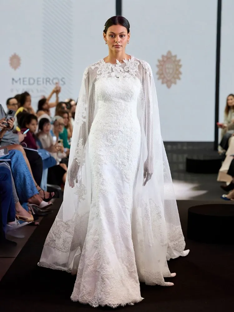 Mulher de pele clara usando vestido de noiva em desfile, com capa, rendas e bordados