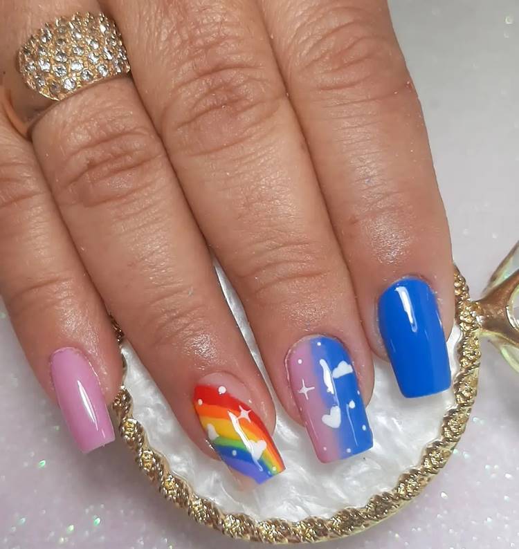 Mão com unhas decoradas coloridas: Polegar: não aparece na foto Indicador: azul Médio: rosa e azul mesclado com corações branco Anelar: arco-íris com corações brancos Mínimo rosa