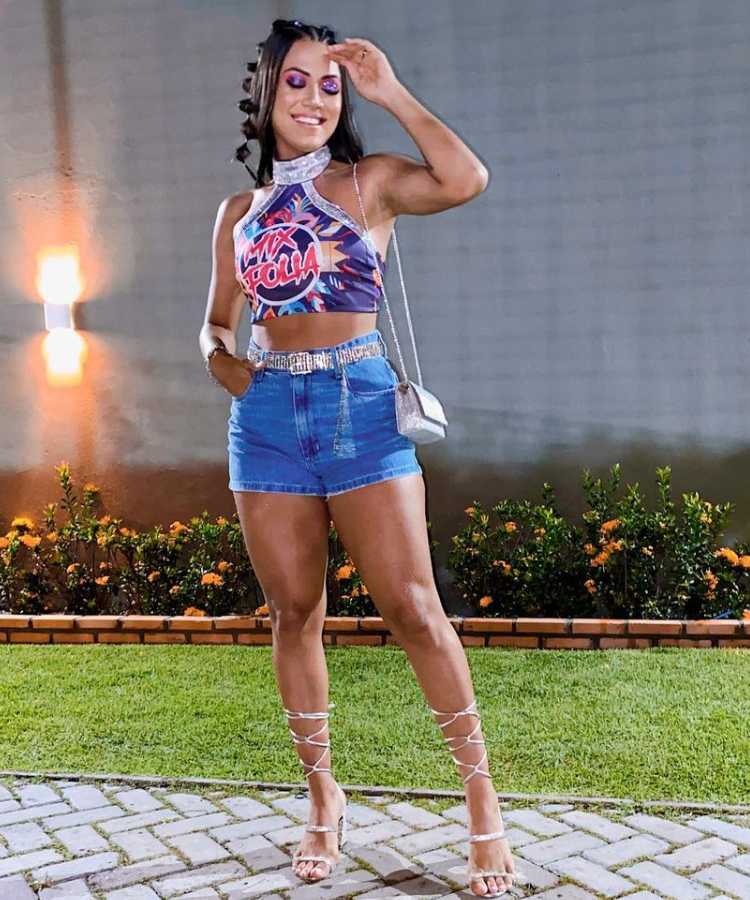 Mulher de pele morena com abadá roxo customizado com paetês nas bordas e gola, além de shorts jeans e bolsa prata