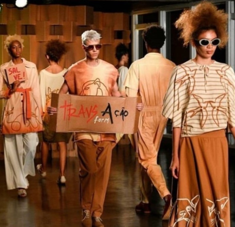 Modelos desfilando para Ronaldo Silvestre com roupas em tom de marrom, branco, laranja e creme, onde se lê uma placa com as palavras TransAção