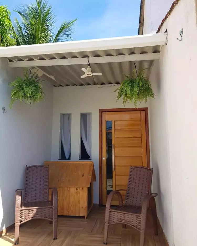 Imagem de fachada de casa simples e pequena. Porta de madeira, samambaia pendurada, telhado de ternit. Casinha de cachorro grande no canto e 2 cadeiras na frente
