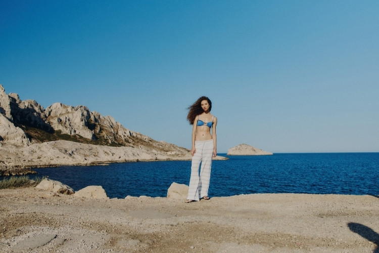 Foto de modelo em praia de Marseille usando top de biquíni azul com calça de crochê branca da coleção verão 2026 da Salinas