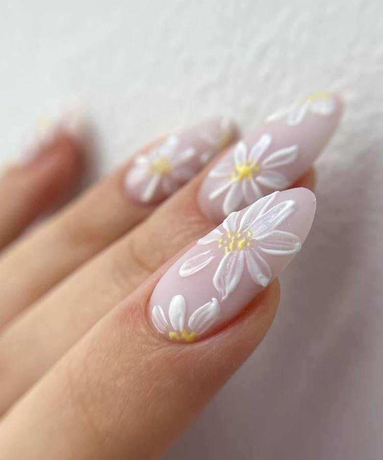Unhas em formato amendoado decoradas com esmalte nude rosado e desenhos delicados de margaridas brancas com miolo amarelo. O acabamento minimalista e artístico destaca sofisticação e frescor. Fundo branco com textura suave complementa a elegância do design floral.






