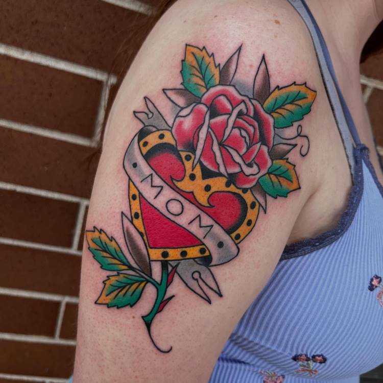 A imagem mostra uma tatuagem colorida no braço de uma pessoa, no estilo tradicional. Ela apresenta um coração vermelho e amarelo, perfurado por um punhal e com uma faixa cinza escrito "MOM". Uma rosa vermelha e folhas verdes e amarelas adornam o desenho.







