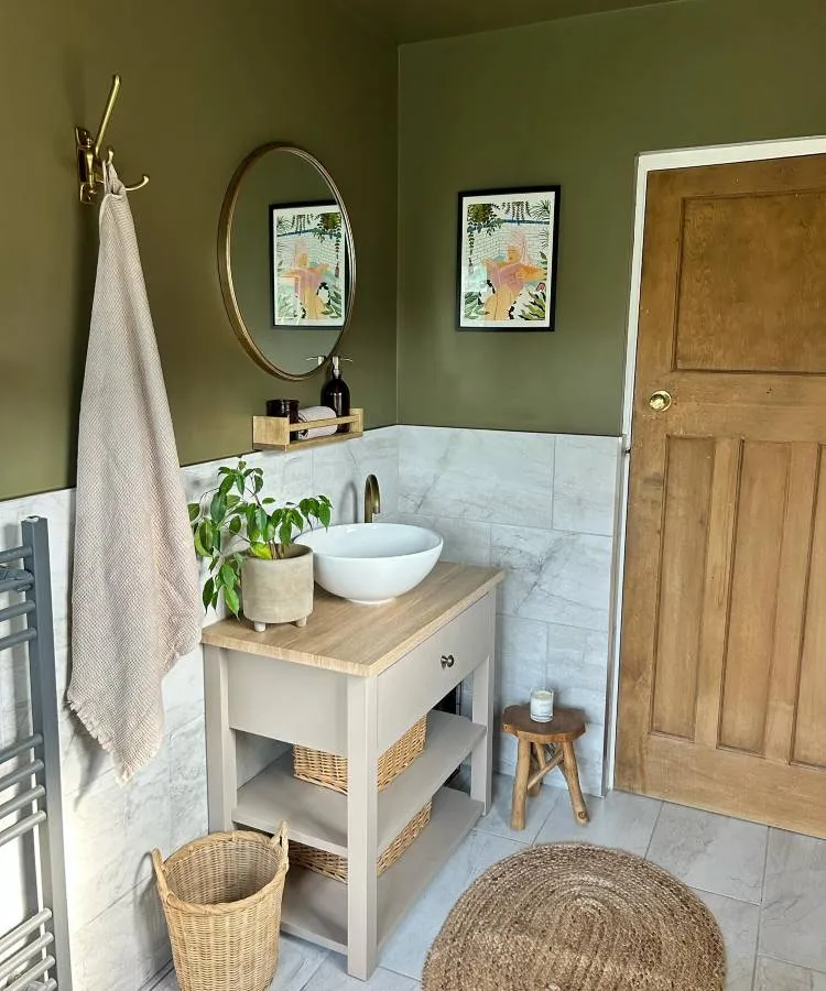 Um banheiro com paredes verde-oliva e azulejos de mármore branco. Possui uma pia de bancada branca sobre um gabinete bege com prateleiras de vime, um espelho redondo dourado e uma porta de madeira. Plantas e cestos de vime complementam a decoração rústica.

