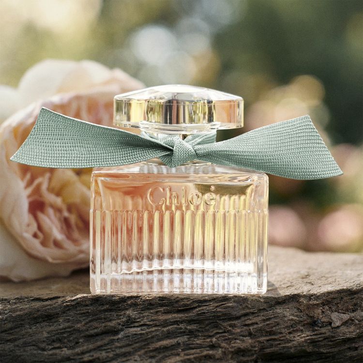 Um frasco de perfume "Chloé" transparente e plissado, com um elegante laço verde-água. A imagem transmite frescor e delicadeza, com uma flor em tons suaves ao fundo, realçando a naturalidade da fragrância- perfumes femininos amadeirados.
