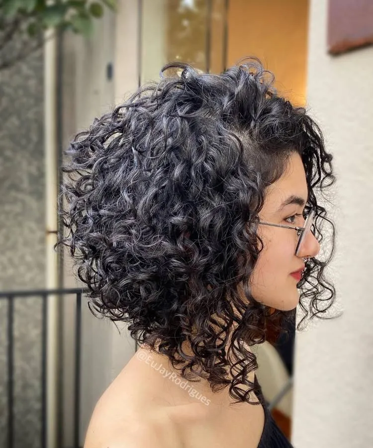 Uma mulher de perfil com um corte de cabelo cacheado bob, com cachos escuros e volumosos. Ela usa óculos e batom vermelho, e uma blusa preta, com a pele clara.