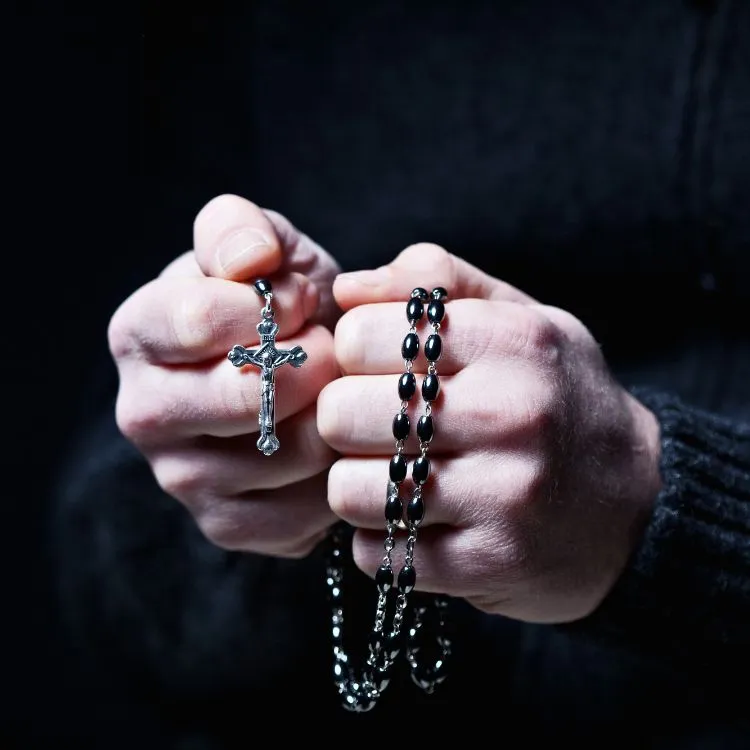A imagem mostra duas mãos segurando firmemente um terço preto com contas brilhantes e uma cruz de prata no final. As mãos estão em foco contra um fundo escuro