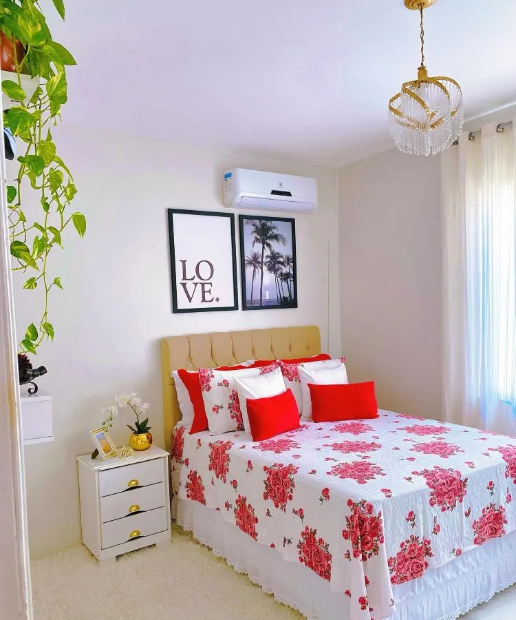 A imagem mostra uma decoração fácil para quarto com paredes brancas, cama de casal com roupa de cama branca estampada com flores vermelhas, um quadro preto e branco com a palavra “LOVE” e uma imagem de palmeiras acima da cama. Ao lado da cama, há um criado-mudo branco com uma luminária e livros empilhados, além de uma planta verde pendurada no teto ao lado de um lustre dourado.

