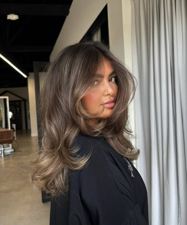 A imagem exibe uma mulher com cabelo castanho iluminado, volumoso e com ondas leves que emolduram o rosto. Ela veste uma blusa preta e olha de lado, exibindo maquiagem natural. O fundo é um salão de beleza com cortinas claras e luzes.







