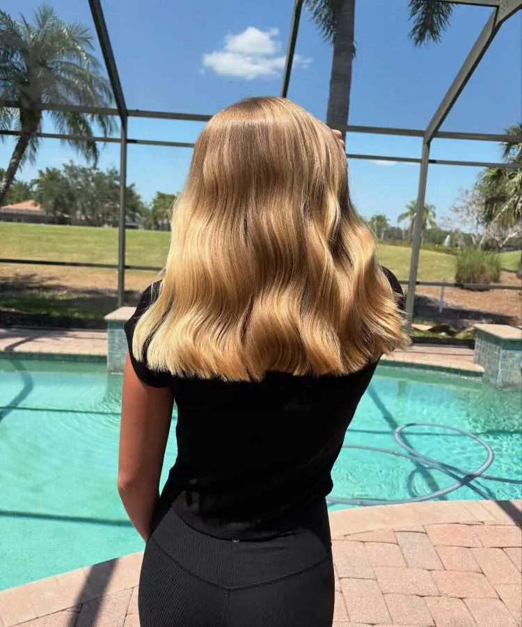 Mulher de costas com cabelos loiros longos, em ondas largas e brilhantes, destacando um corte blunt com pontas retas. Ela veste blusa preta enquanto está em um ambiente externo ensolarado, próximo a uma piscina com gramado ao fundo.