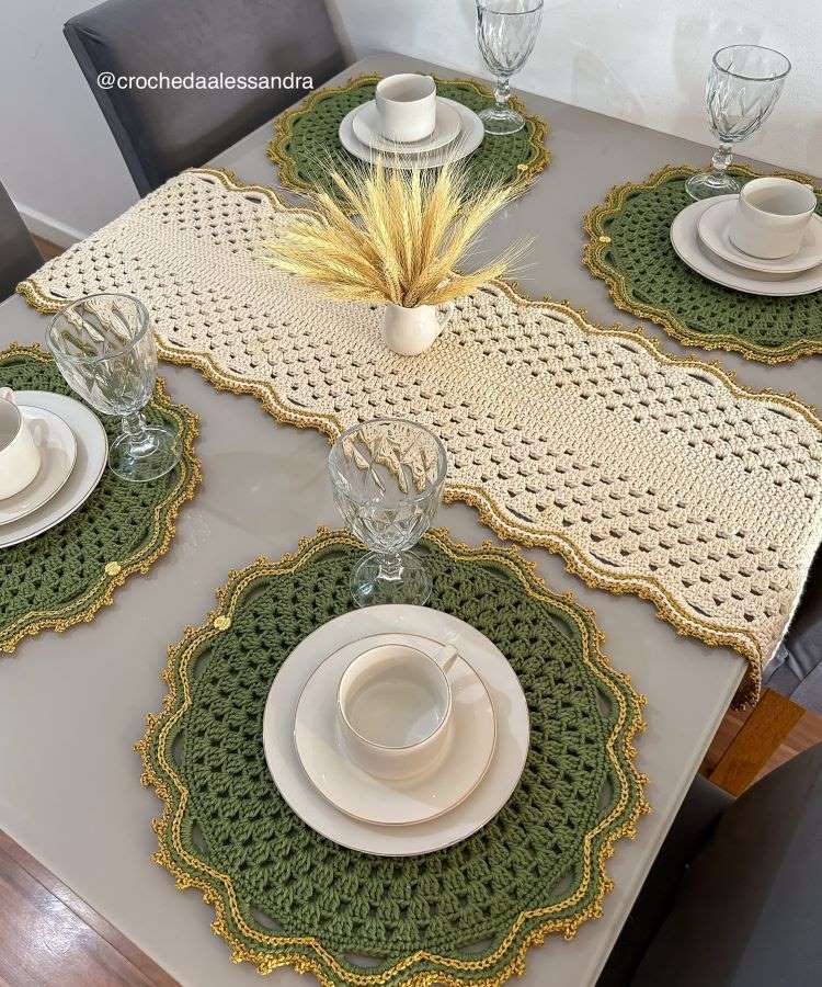 Mesa elegante com sousplats de crochê verde e bordas douradas. Louças brancas e taças de vidro complementam a decoração. Um arranjo central de trigo em vaso branco completa o visual sofisticado.